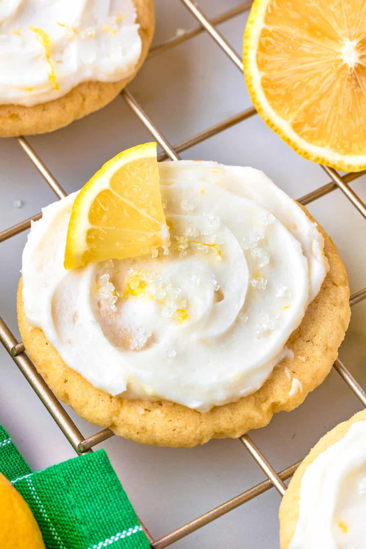 Lemon Sugar Cookies A frosted lemon cookie topped with a lemon wedge and sugar crystals sits on a cooling rack, with a sliced lemon and part of a green towel nearby.