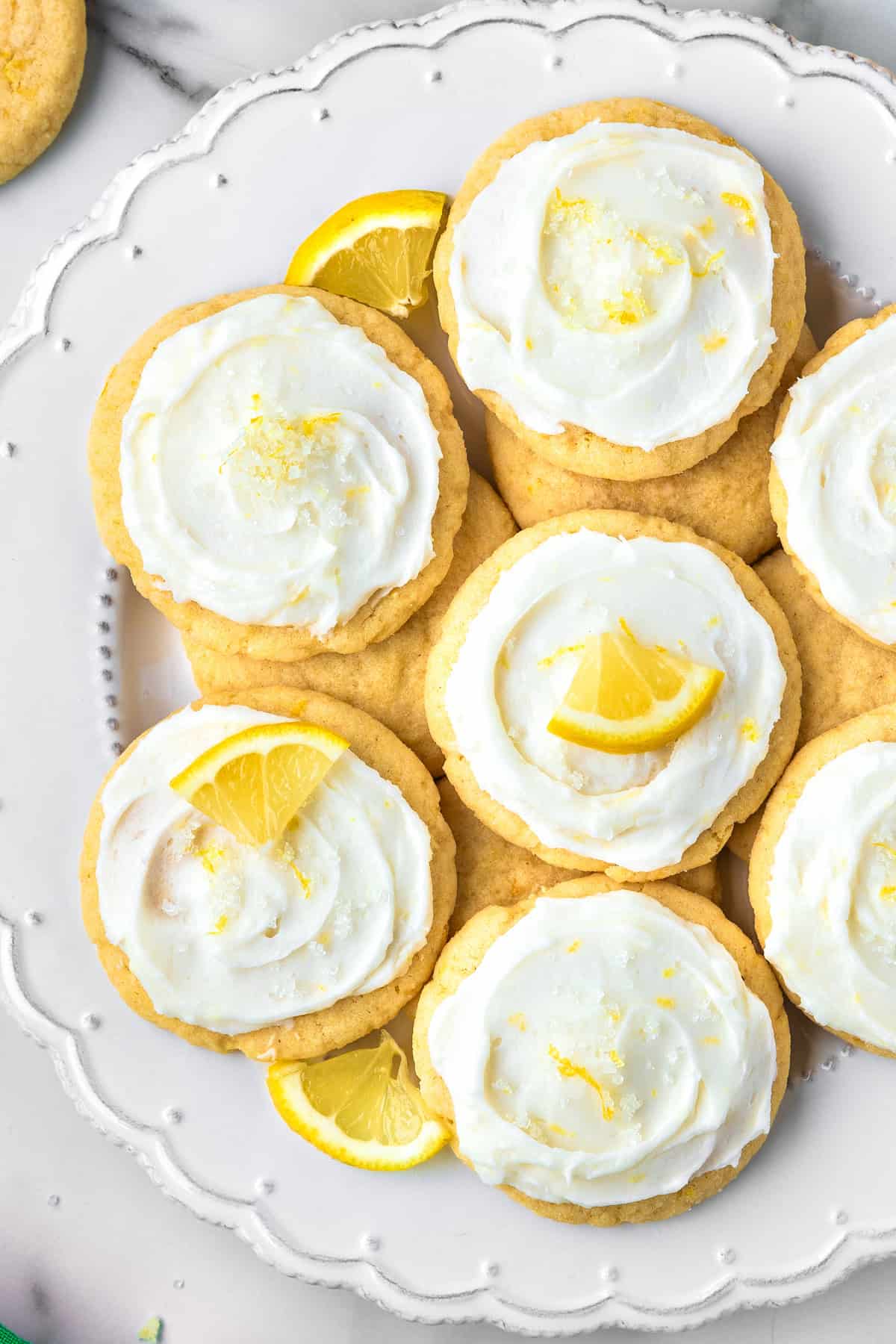 Lemon Sugar Cookies A plate of lemon cookies with white frosting, garnished with lemon zest and small lemon wedges, arranged on a decorative white plate.