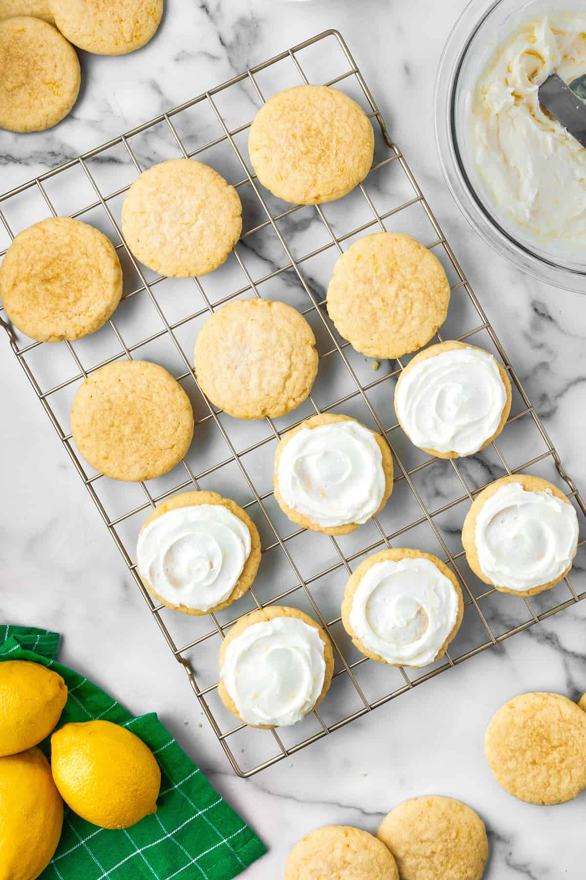 Lemon Sugar Cookies Round lemon cookies cool on a wire rack, some with white frosting. A green towel with lemons and a bowl of frosting are nearby on a marble countertop.