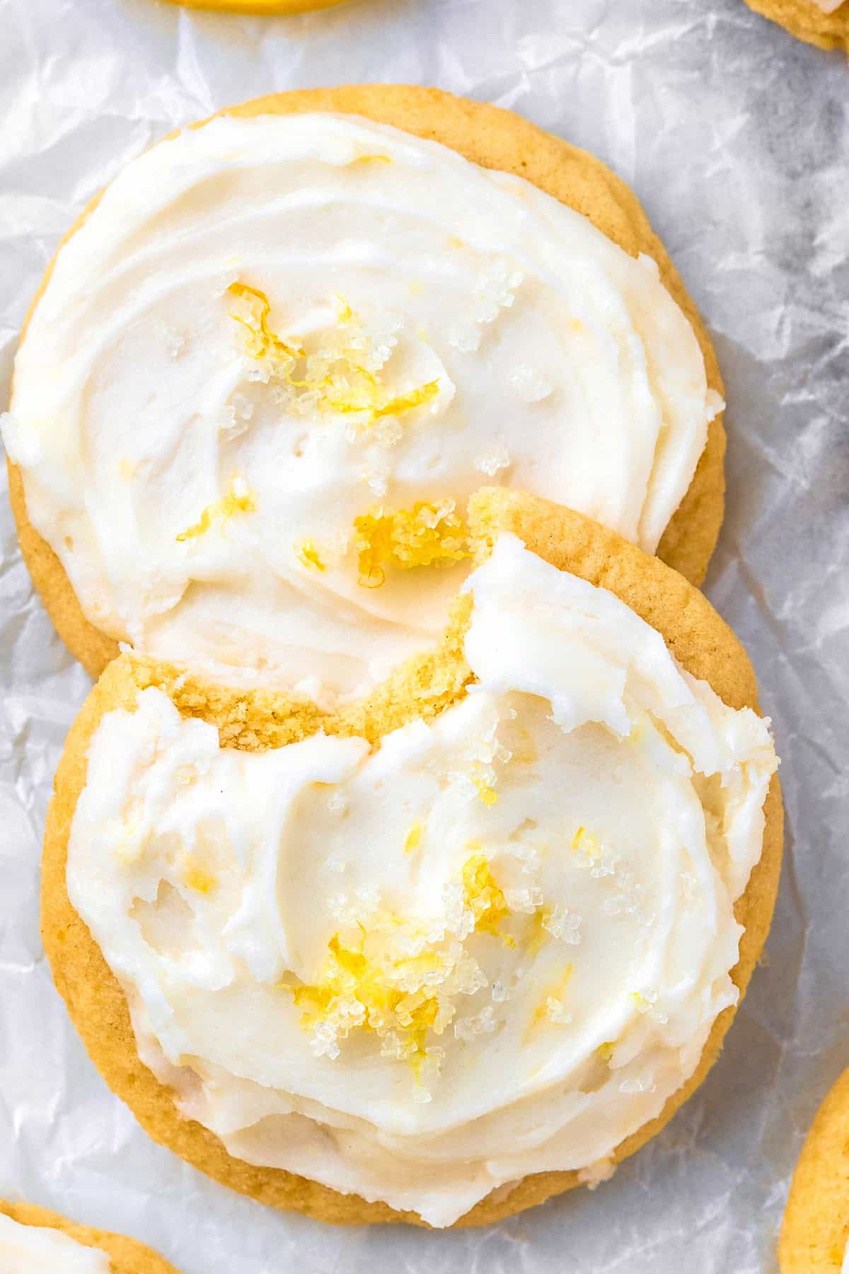 Lemon Sugar Cookies Two round cookies with creamy white frosting and lemon zest on top, placed on crinkled parchment paper. One cookie overlaps the other.