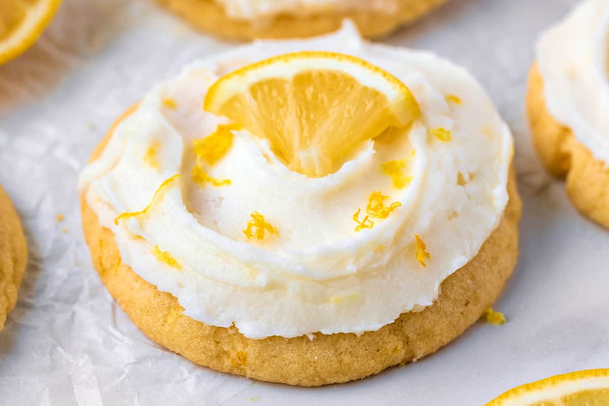 Lemon Sugar Cookies A round sugar cookie with white frosting, sprinkled with lemon zest, and topped with a small lemon wedge, resting on white parchment paper.