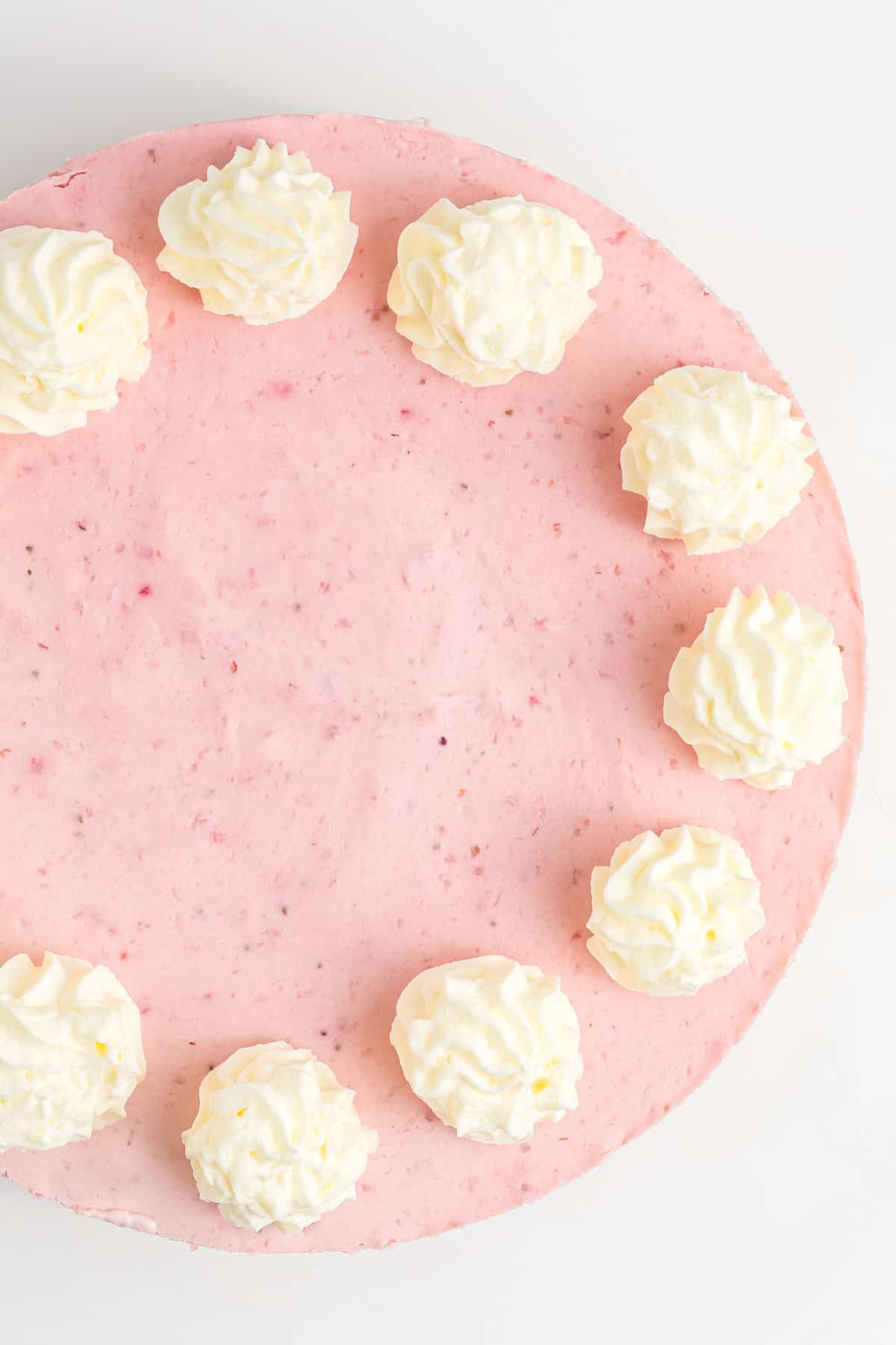 A round pink cheesecake topped with swirls of whipped cream arranged in a semi-circle on a white background.