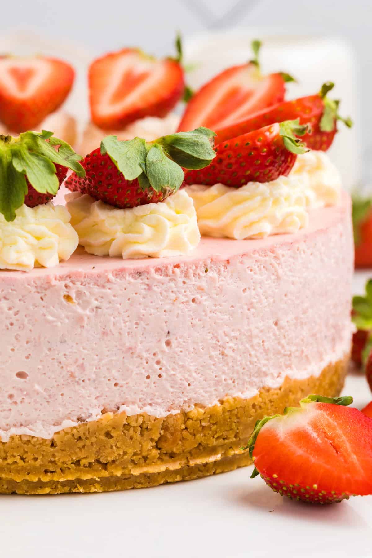 A close-up of a strawberry cheesecake with a crumbly golden crust, creamy pink filling, whipped cream swirls, and fresh strawberry halves on top.