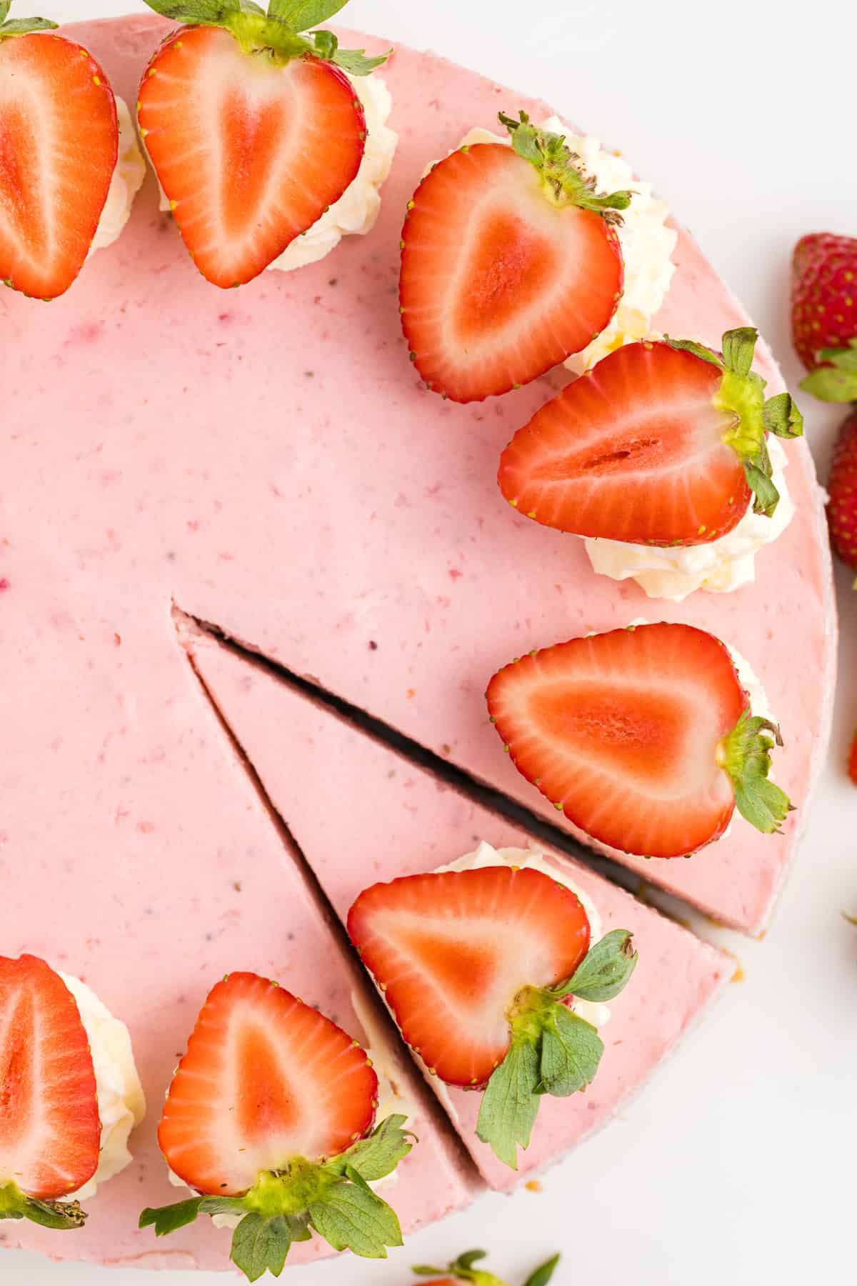 A round strawberry cheesecake with a slice cut out, topped with fresh strawberry halves and dollops of whipped cream. 