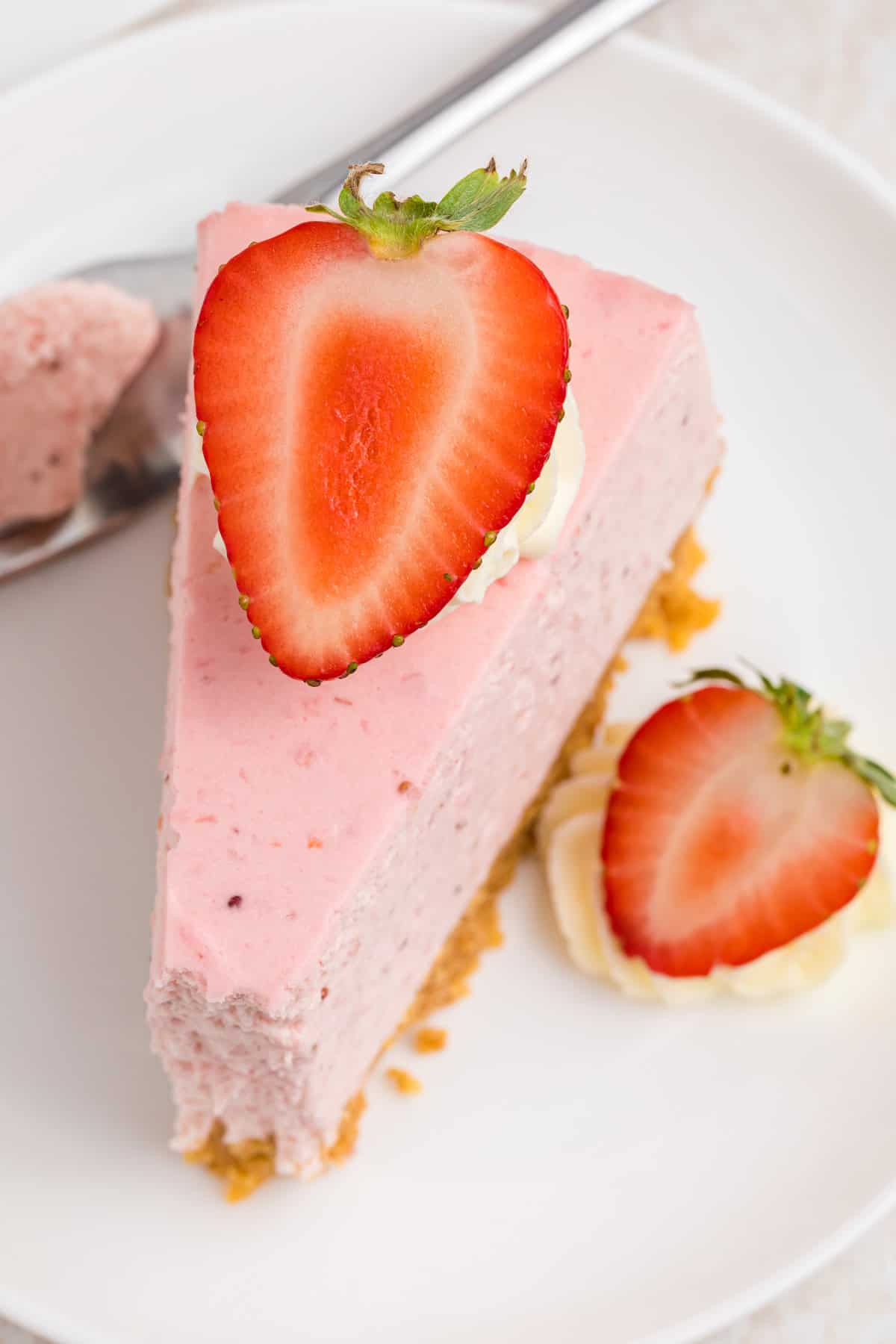 A slice of strawberry cheesecake with a graham cracker crust, topped with whipped cream and a halved fresh strawberry.