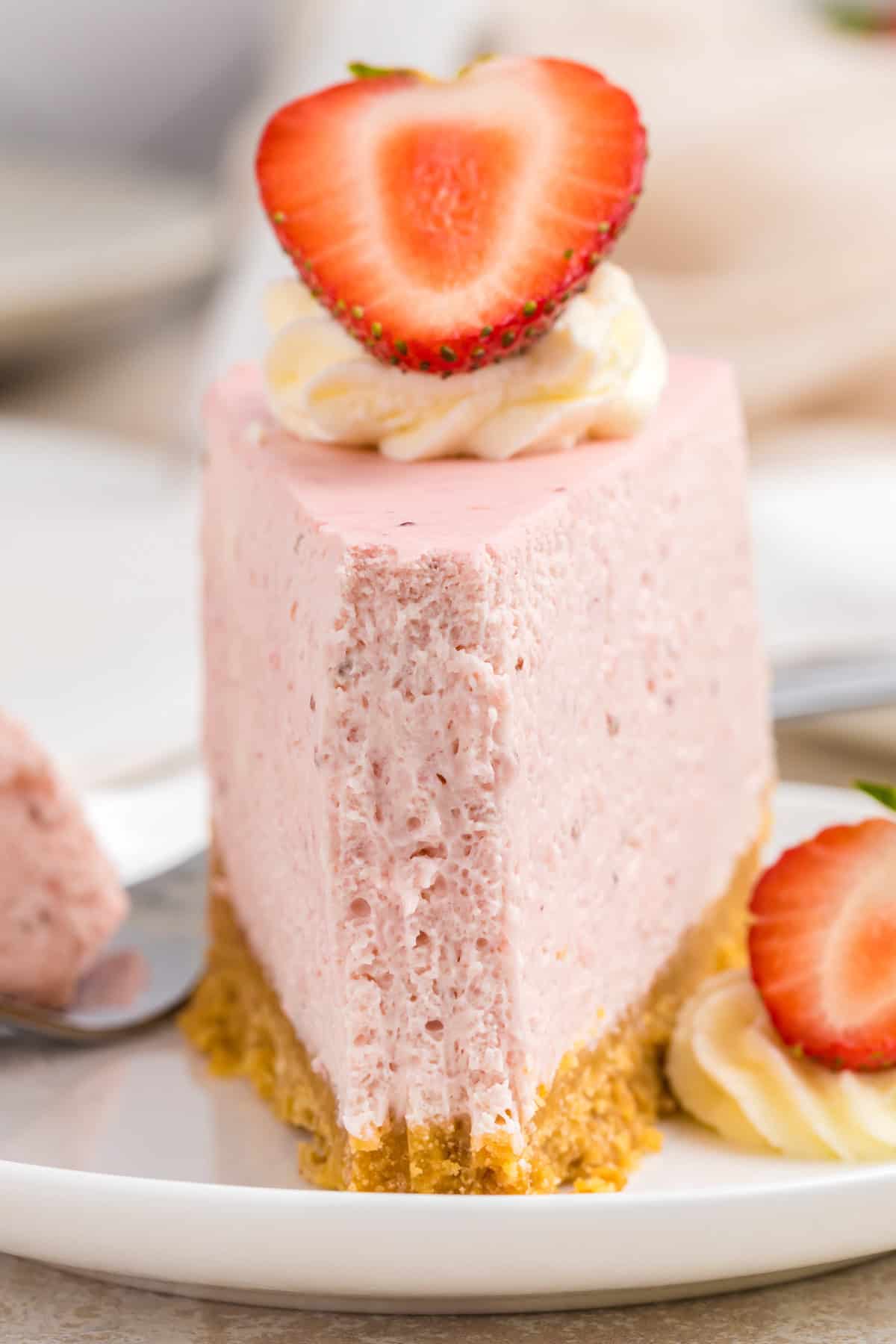A slice of creamy pink strawberry cheesecake with a crumbly crust, topped with whipped cream and a halved strawberry.