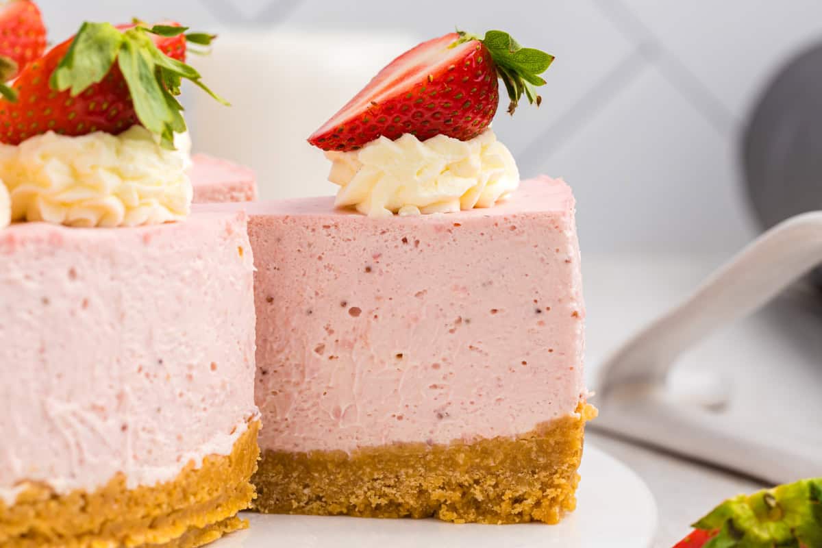 A slice of strawberry cheesecake with a graham cracker crust is topped with whipped cream and half a fresh strawberry.