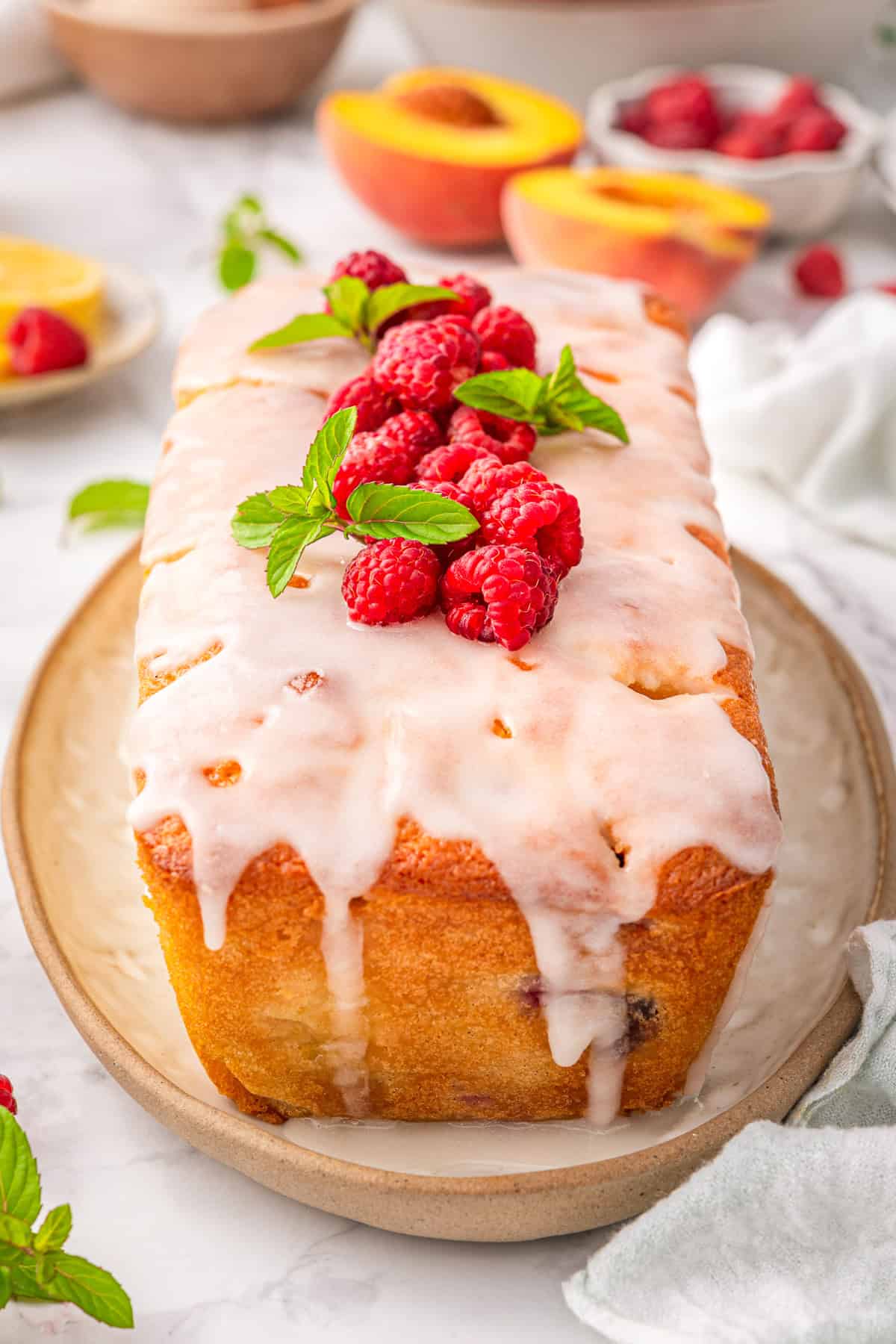 Raspberry Peach Loaf with Lemon Glaze A loaf cake topped with white icing, fresh raspberries, and mint leaves sits on a beige plate.