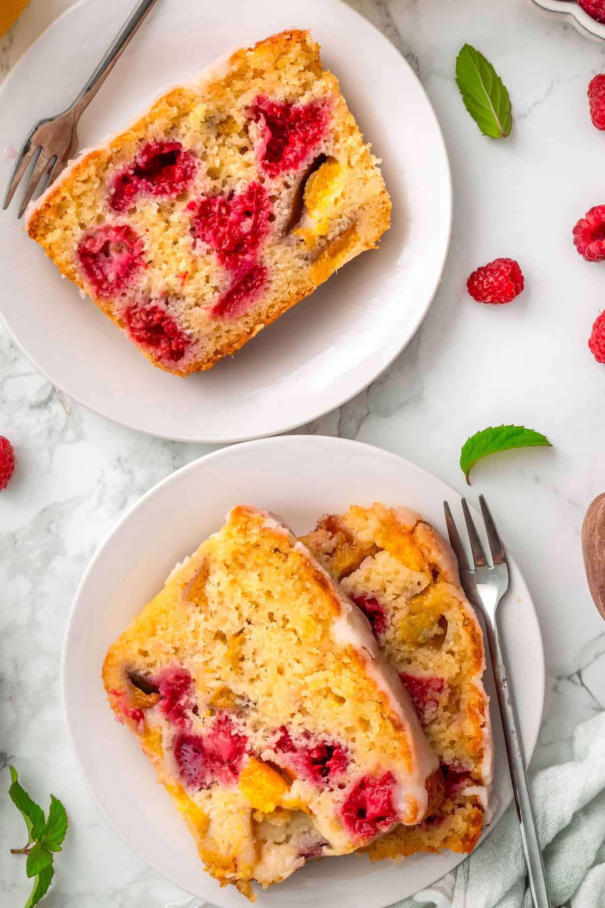 Raspberry Peach Loaf with Lemon Glaze Two white plates with slices of fruit-studded pound cake, featuring visible raspberries and yellow fruit, garnished with scattered fresh raspberries and mint leaves on a marble surface.