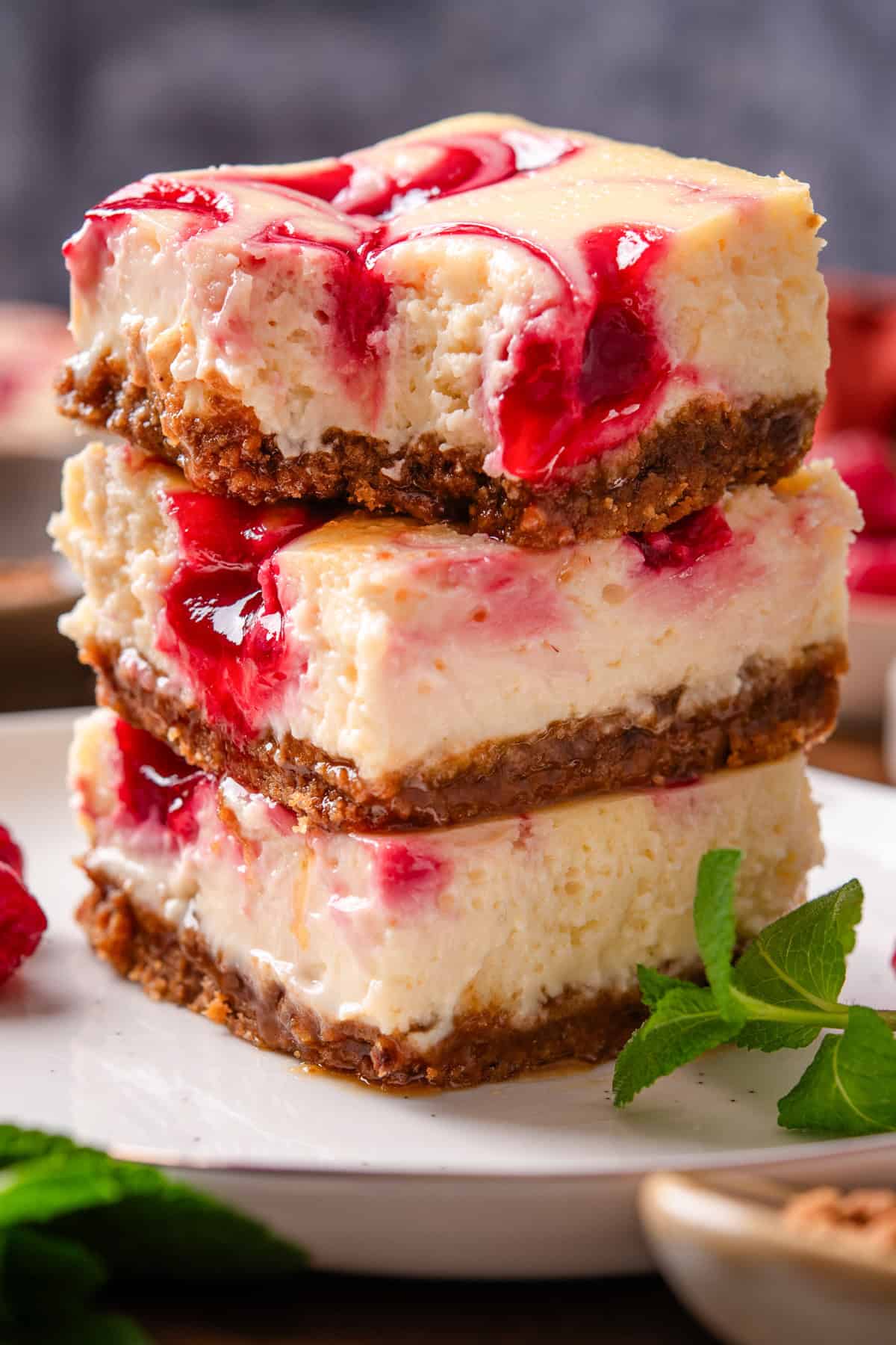 Three cheesecake bars with a graham cracker crust and raspberry swirl are stacked on a white plate.