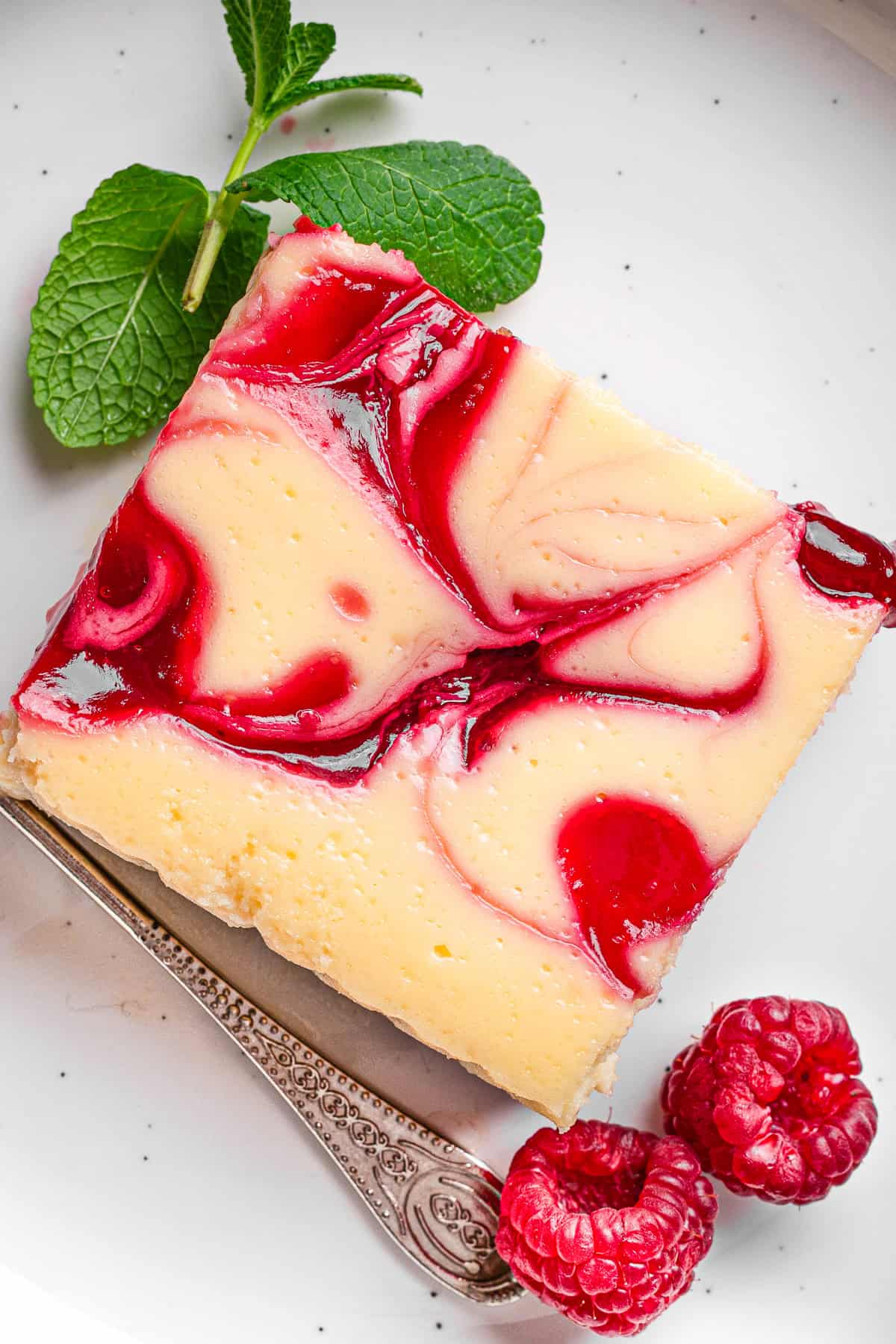 A square slice of cheesecake bar with raspberry swirl, garnished with a mint sprig and fresh raspberries.