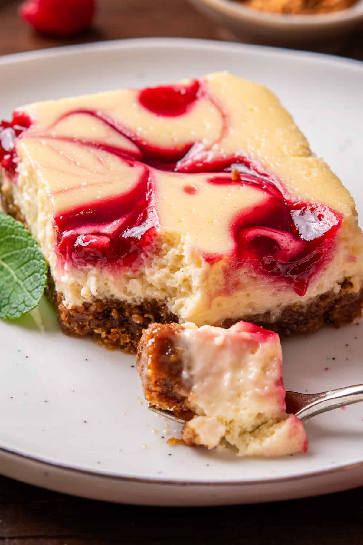 A slice of cheesecake bar with a graham cracker crust and raspberry swirl, garnished with a mint leaf.