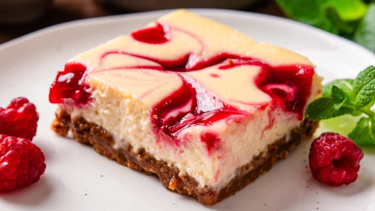 A slice of raspberry swirl cheesecake with a graham cracker crust sits on a white plate.