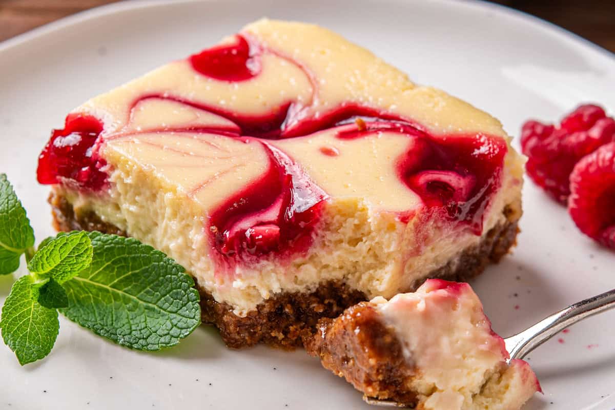 A creamy cheesecake bar with a raspberry swirl on a crumbly crust sits on a white plate.
