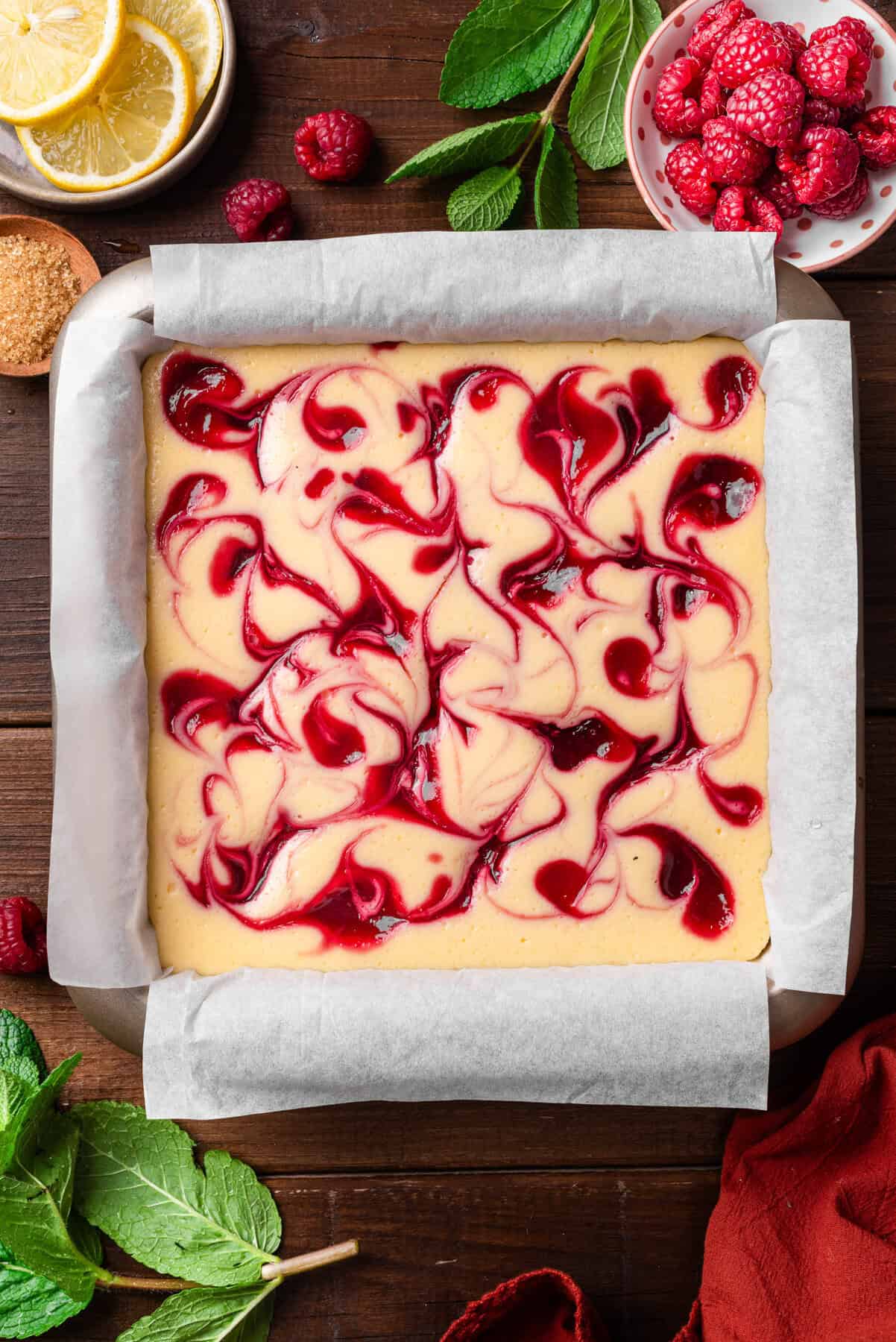 A square baking pan lined with parchment paper holds unbaked cheesecake swirled with raspberry sauce.  