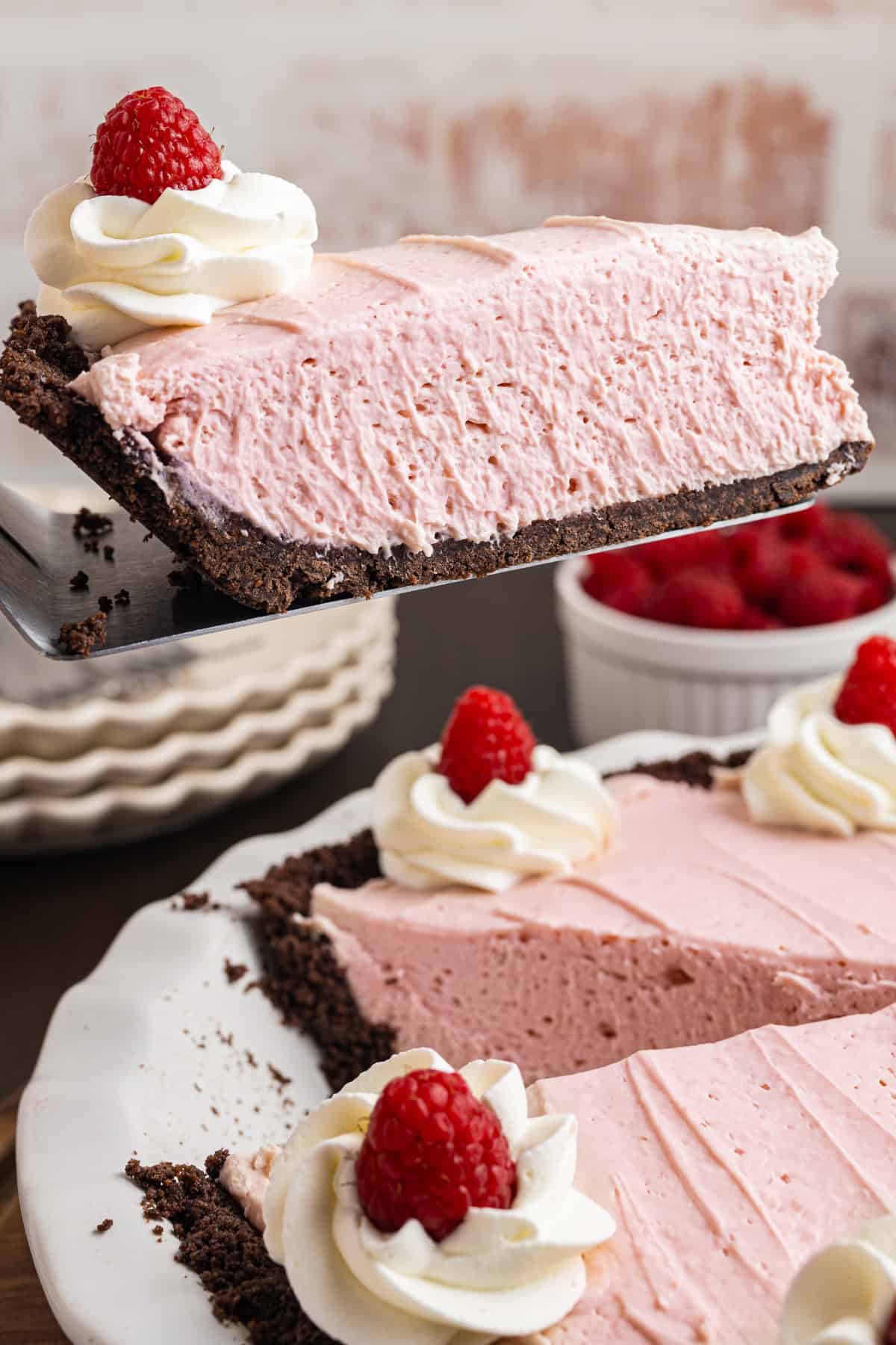 A slice of pink raspberry cream pie with chocolate crust is being lifted from a pie dish. The pie is topped with whipped cream and raspberries. 