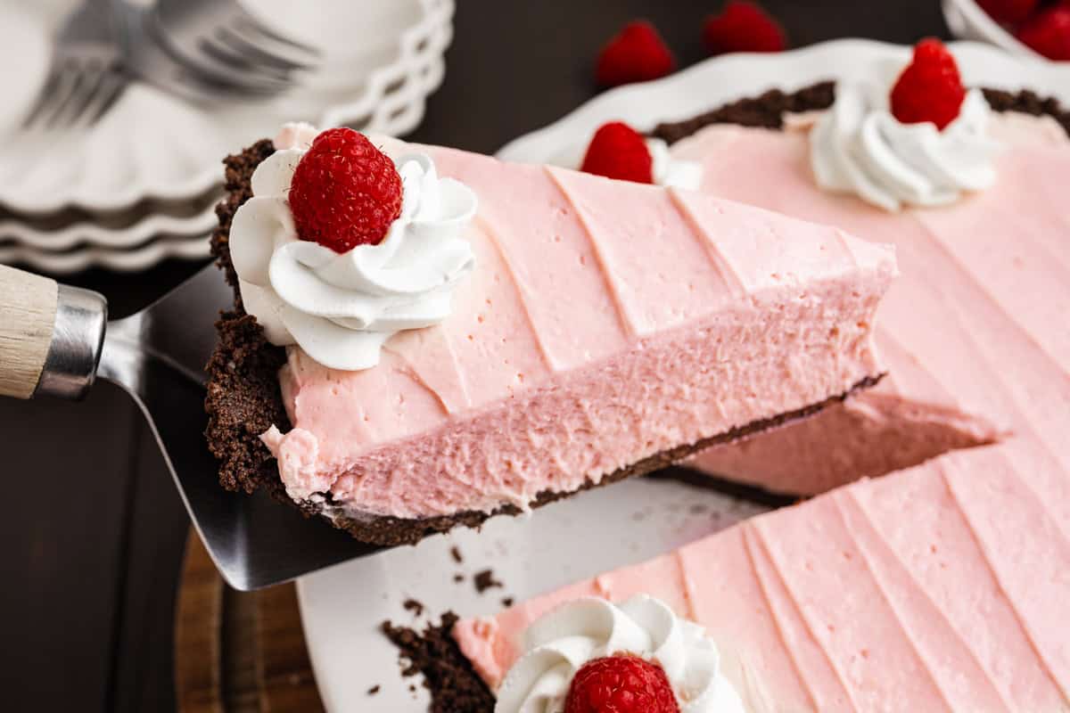 A slice of pink raspberry cream pie with a chocolate crust is lifted from a pie dish, topped with whipped cream and a raspberry. 