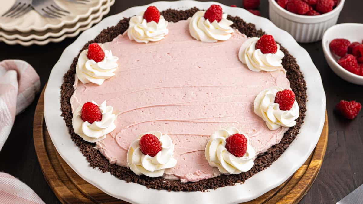 A pink raspberry pie with a chocolate crust, topped with swirls of whipped cream and fresh raspberries, sits on a white pie dish. 