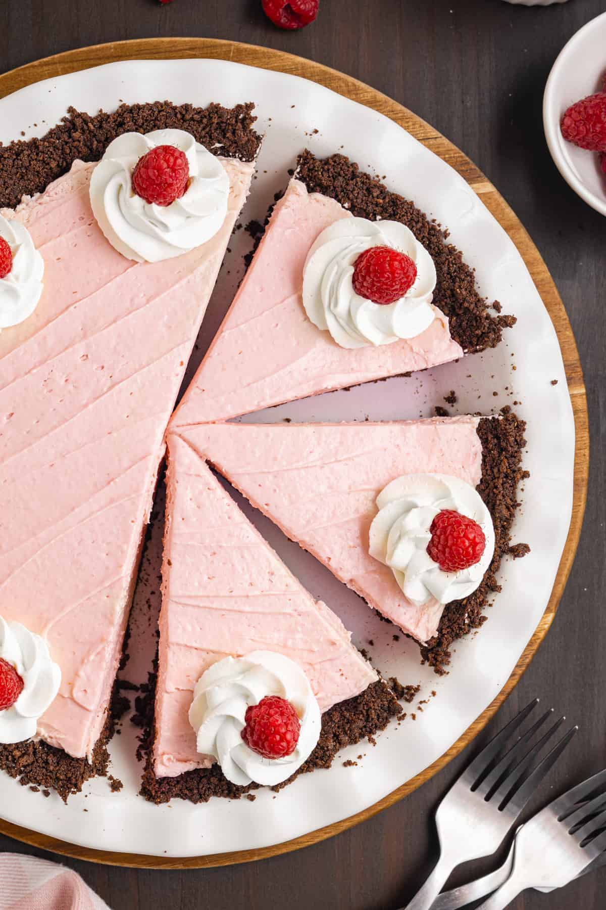 A pink raspberry cream pie with a chocolate crust, topped with whipped cream and raspberries, sits on a white plate. 
