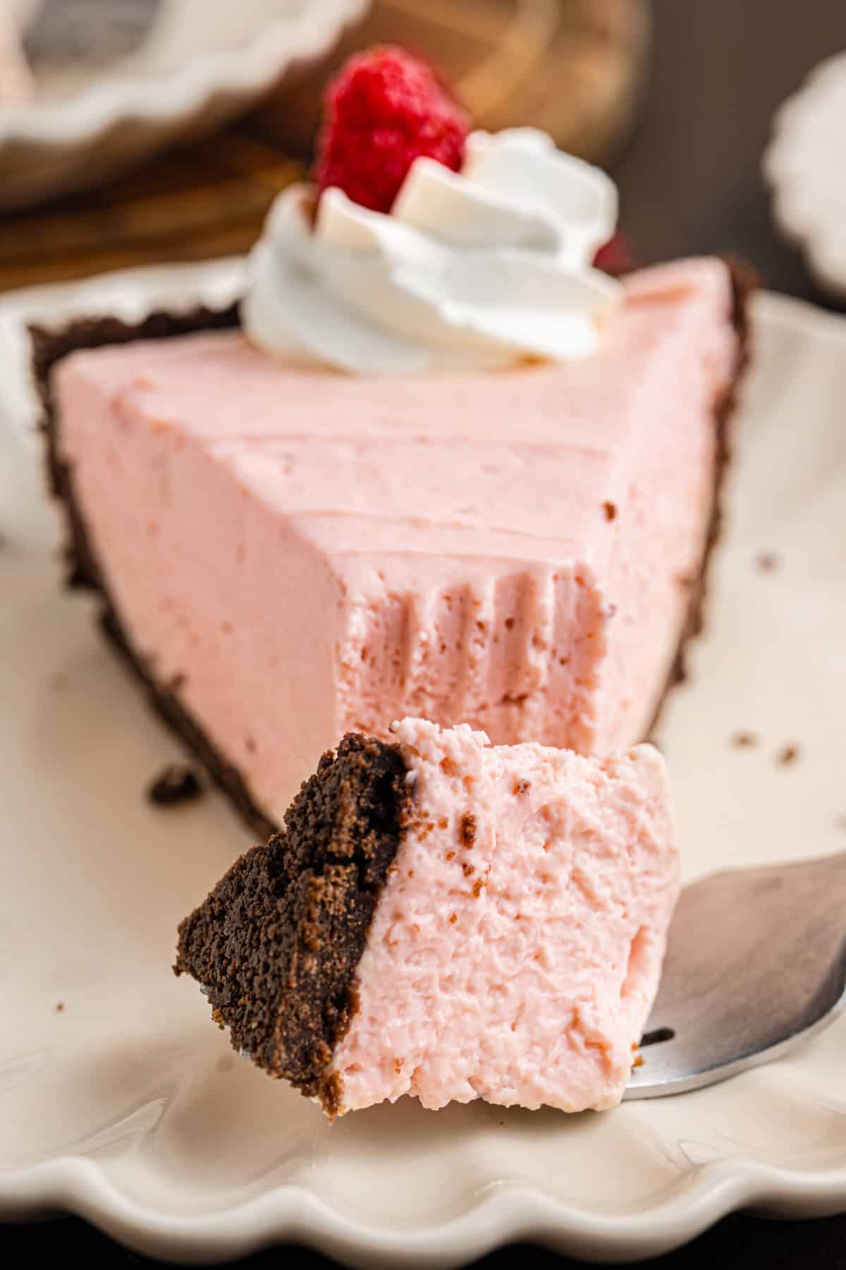 A slice of pink raspberry cream pie with a chocolate crust, topped with whipped cream and a raspberry. 