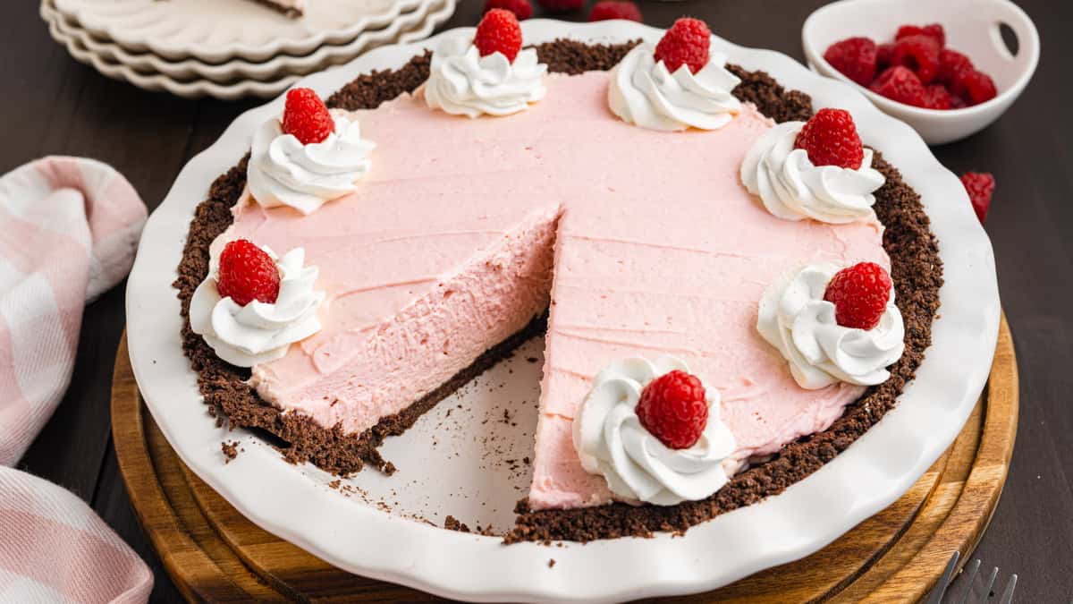 A pink raspberry cream pie in a chocolate crust sits on a white plate. The pie is topped with whipped cream and raspberries, with one slice missing. 