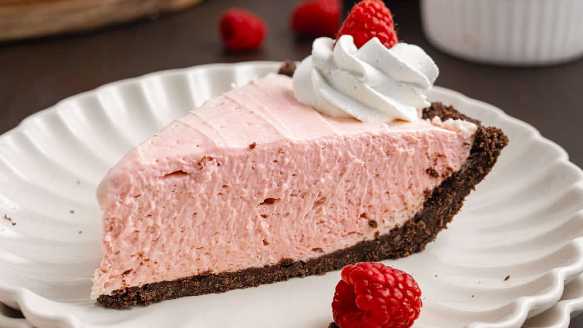 A slice of pink raspberry cheesecake with a chocolate crust, topped with whipped cream and served on a white plate.