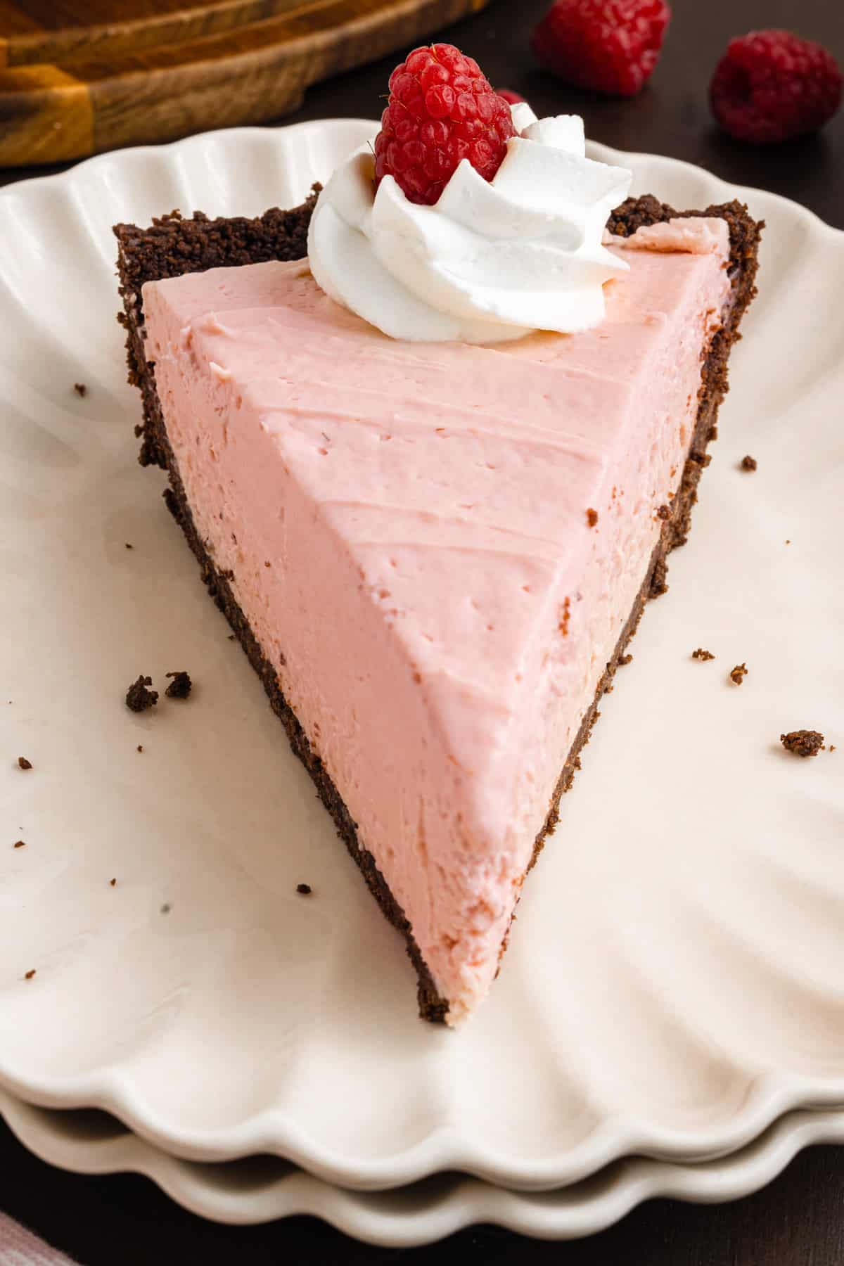 A slice of pink raspberry cream pie with a chocolate crust sits on a scalloped white plate, topped with whipped cream and a fresh raspberry.