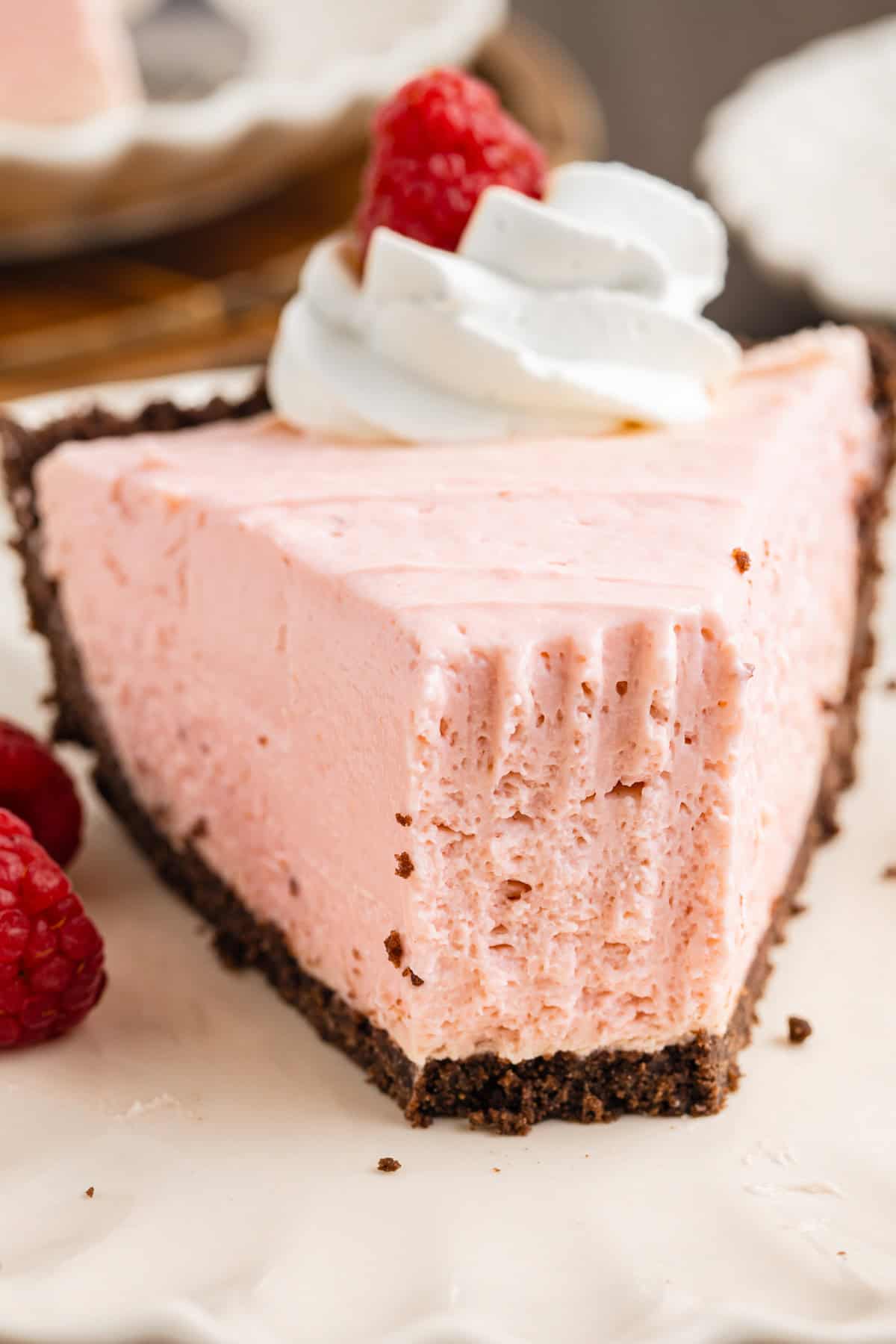 A slice of creamy pink raspberry pie with a chocolate crust, topped with whipped cream and a raspberry. A bite has been taken from the front. 