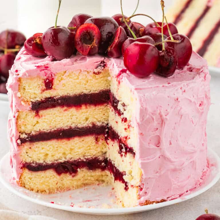 A layered vanilla cake with pink cherry frosting and cherry filling, topped with fresh cherries. A slice has been cut out, showing the cake's layers.