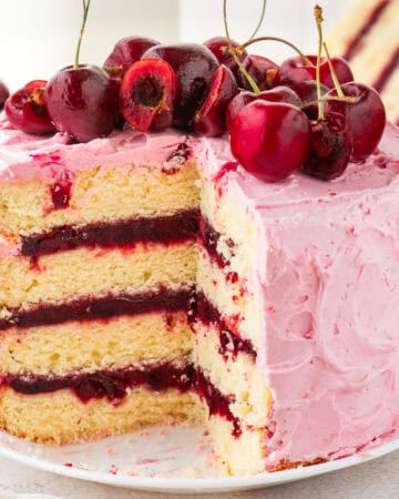 A three-layer vanilla cake with pink cherry frosting, cherry filling between layers, and fresh cherries on top. A large slice has been cut out, revealing the cakes texture and vibrant colors.