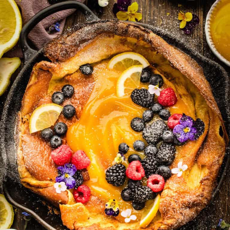 A Dutch baby pancake topped with lemon curd, fresh blueberries, raspberries, blackberries, lemon slices, and edible flowers in a cast-iron skillet.