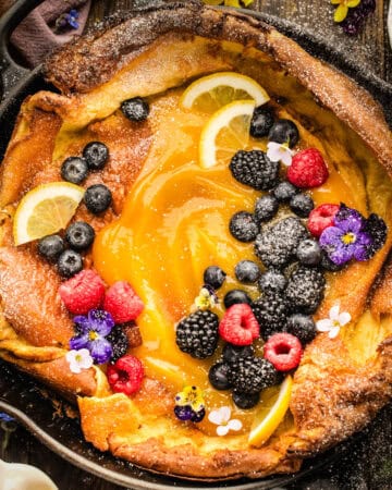 A Dutch baby pancake in a skillet, topped with lemon curd, lemon slices, raspberries, blackberries, blueberries, and edible flowers, dusted with powdered sugar.