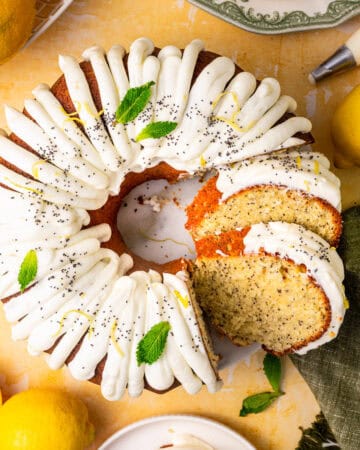 A lemon poppy seed bundt cake with white icing, garnished with poppy seeds, lemon zest, and mint leaves. Slices are cut, showing the cake's texture.