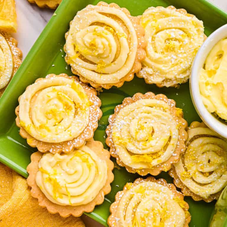 Round lemon shortbread cookies topped with swirled buttercream and garnished with lemon zest and powdered sugar, are arranged on a green plate.