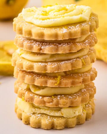 A stack of lemon shortbread cookies with creamy filling and powdered sugar dusting sits on a light surface. A whole lemon and yellow cloth are blurred in the background.