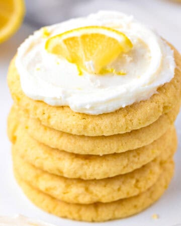 A stack of frosted lemon sugar cookies topped with a lemon wedge, with a fresh lemon in the background.