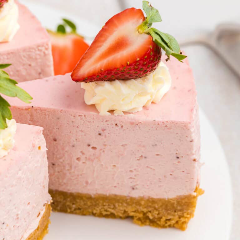 A slice of pink strawberry cheesecake with a crumbly golden crust, topped with whipped cream and half a fresh strawberry on top.