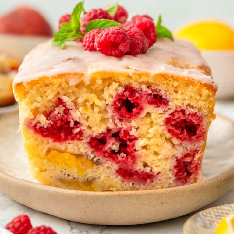 A thick slice of raspberry lemon loaf cake with glaze sits on a plate, topped with fresh raspberries and mint. The cake has visible raspberries baked inside and a moist, yellow crumb.