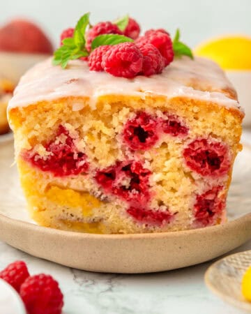 A sliced loaf of raspberry and peach bread with white icing, garnished with fresh raspberries and mint, sits on a beige plate with fruit in the background.
