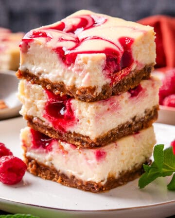 Three raspberry swirl cheesecake bars are stacked on a plate. The bars have a creamy texture, graham cracker crust, and vibrant red raspberry swirls on top.