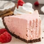 A slice of pink cheesecake with a chocolate crust, topped with whipped cream and a raspberry.