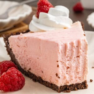 A slice of pink cheesecake with a chocolate crust, topped with whipped cream and a raspberry.