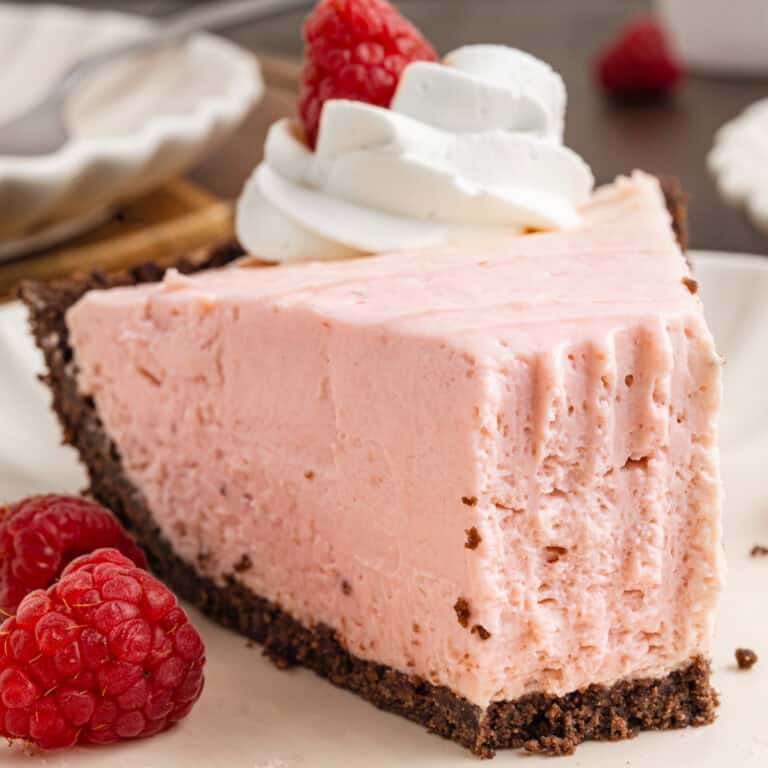 A slice of pink cheesecake with a chocolate crust, topped with whipped cream and a raspberry.