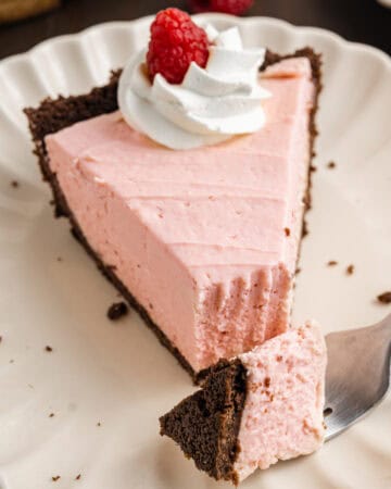 A slice of pink cream pie with a chocolate crust, topped with whipped cream and a raspberry, sits on a white plate.
