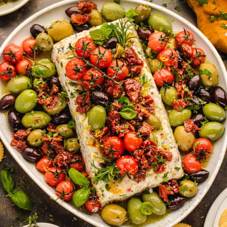 A rectangular block of cheese topped with sun-dried tomatoes, cherry tomatoes, and fresh herbs.
