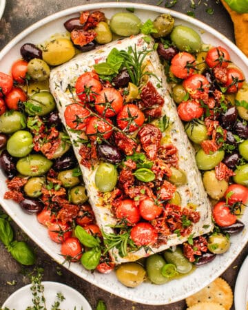 A platter with herbed cheese topped with tomatoes, sun-dried tomatoes, fresh basil, and surrounded by assorted green and black olives.