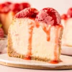 A close-up of a slice of cheesecake topped with whole strawberries and strawberry sauce, with creamy filling and a crumbly crust on a white plate.