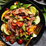 Colorful Thai beef salad with sliced steak, tomatoes, cucumber, bell peppers, fresh herbs, lime wedges, and red chili.