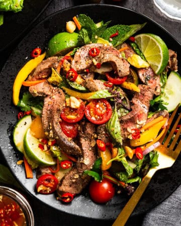 A colorful plate of sliced beef steak salad with lime wedges, cherry tomatoes, cucumber, red chili, bell peppers, avocado, fresh herbs, and a sprinkling of nuts, served with a gold fork.