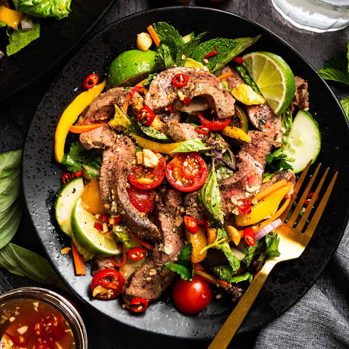 A colorful plate of sliced beef steak salad with lime wedges, cherry tomatoes, cucumber, red chili, bell peppers, avocado, fresh herbs, and a sprinkling of nuts, served with a gold fork.